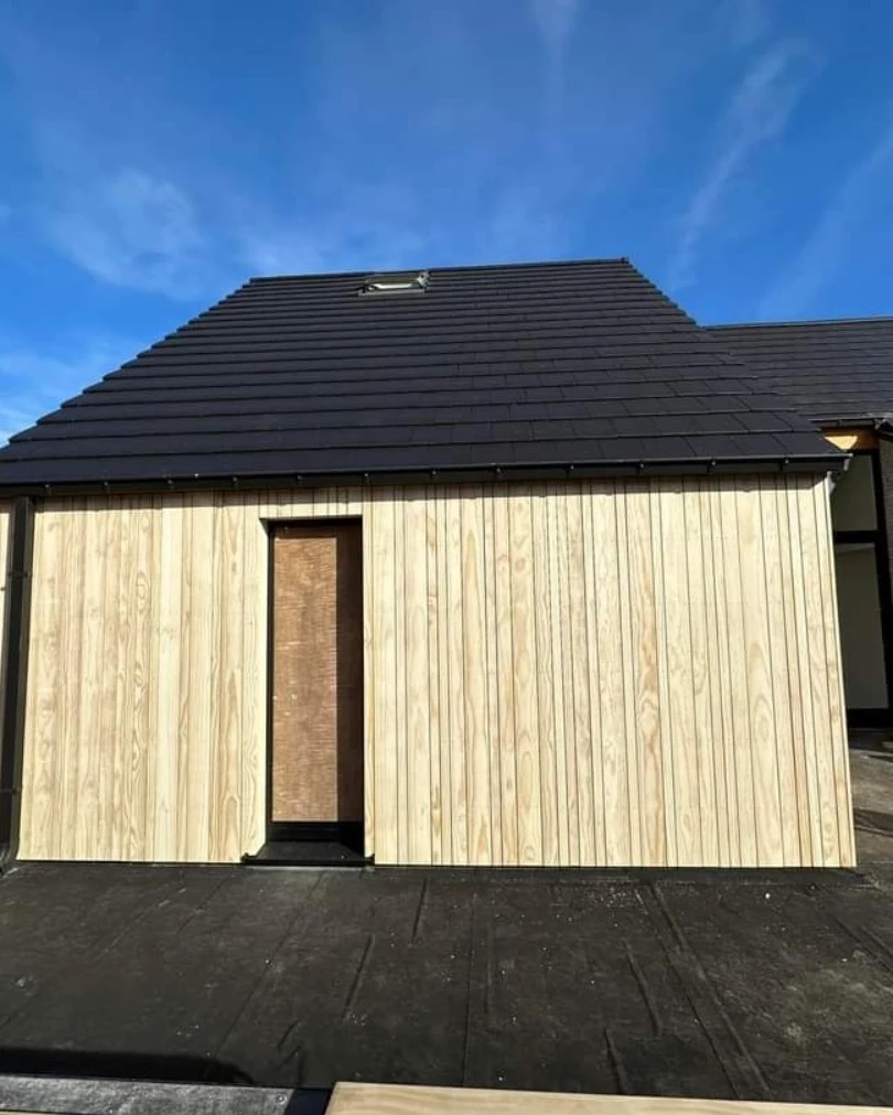 Nowo wybudowany budynek z jasną, pionową okładziną drewnianą i czarnym dachem z dachówką, widoczne okno dachowe i zabezpieczone wejście.