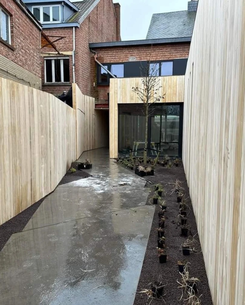 Nowoczesny ogród z drewnianym ogrodzeniem i elementami zabudowy, świeżo po deszczu, z rzędami małych roślin w doniczkach wzdłuż ścieżki.