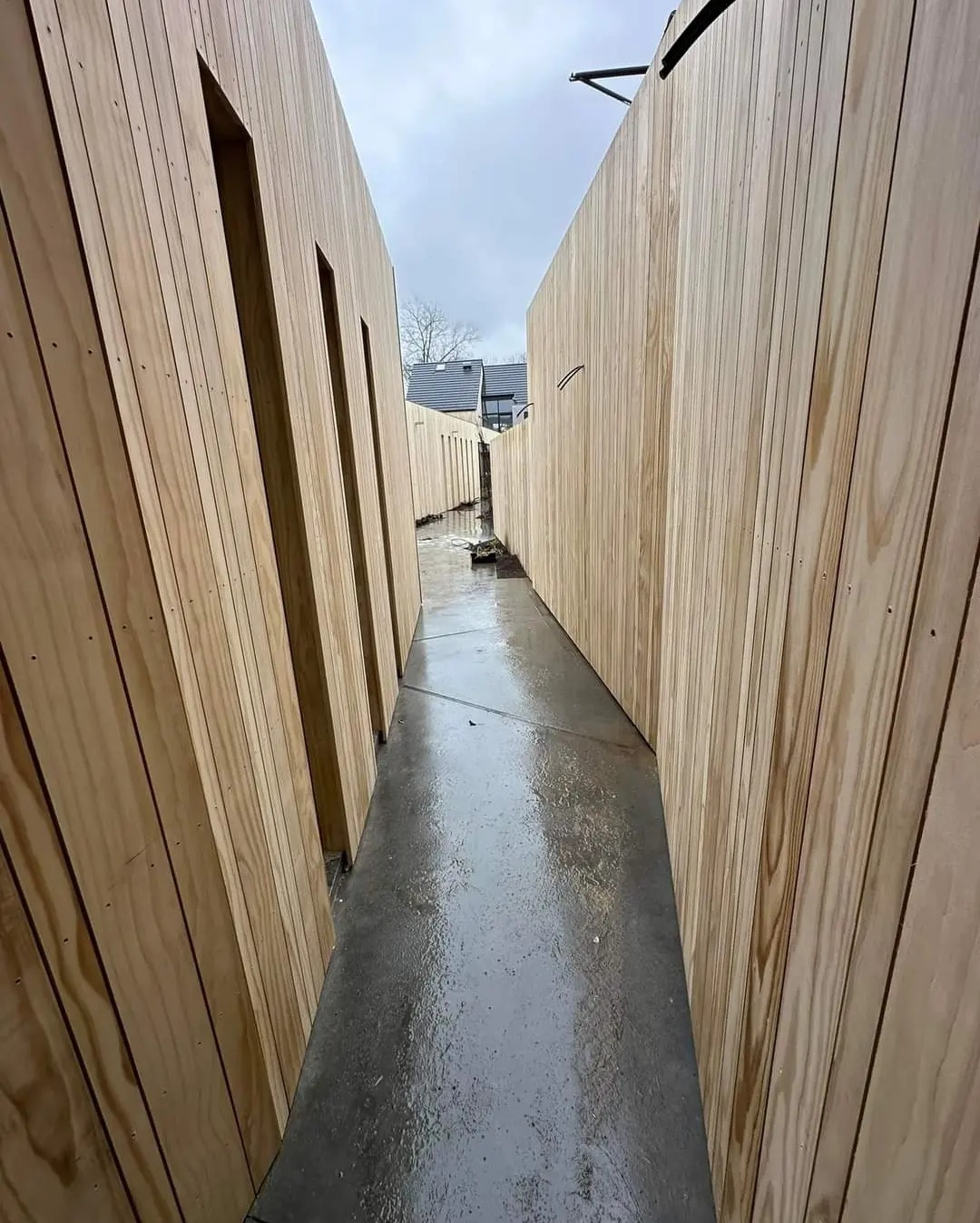 Wąskie przejście między dwiema drewnianymi ścianami z pionowych desek, mokry betonowy chodnik, pochmurne niebo w tle.