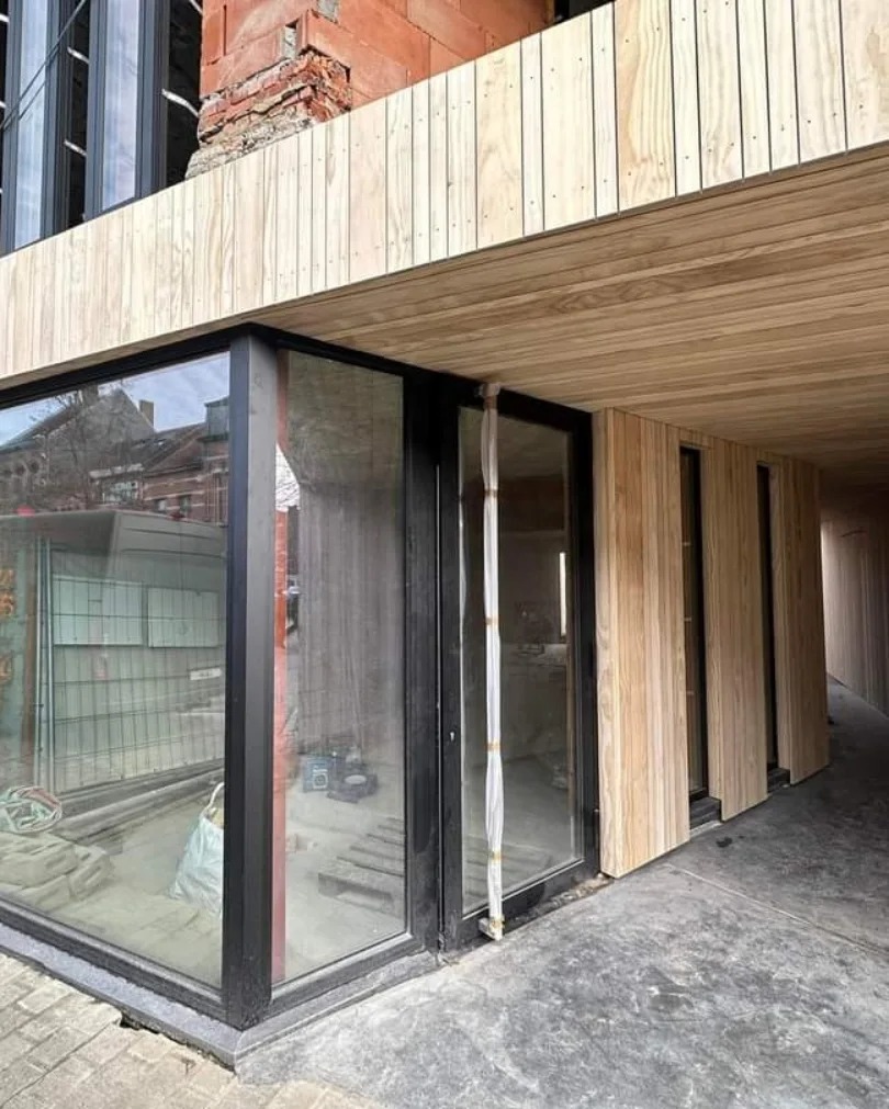 Nowoczesny budynek w trakcie wykończenia, z widocznym drewnianym tarasem i elewacją, surowy mur z cegły w górnej części budynku, duże okna z czarnymi ramami, betonowa podłoga w stanie surowym.