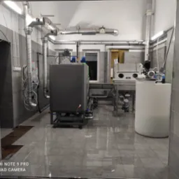 Wnętrze przemysłowe z instalacją rur wentylacyjnych pod sufitem, zbiornikiem i sprzętem do obróbki metali, widoczne płytki podłogowe