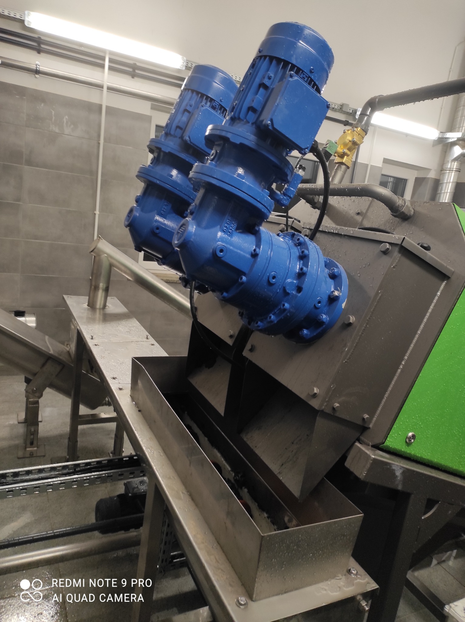 Dwa niebieskie silniki elektryczne z przekładniami, zamontowane na szarej metalowej konstrukcji z elementami ze stali nierdzewnej, prawdopodobnie część większej maszyny przemysłowej, widoczne rury...