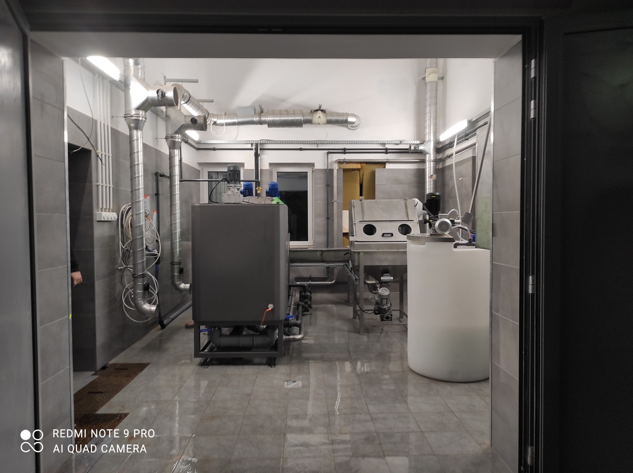 Wnętrze hali produkcyjnej z zainstalowanymi urządzeniami do obróbki metali, widoczne rury wentylacyjne i zbiorniki, szara kolorystyka pomieszczenia.