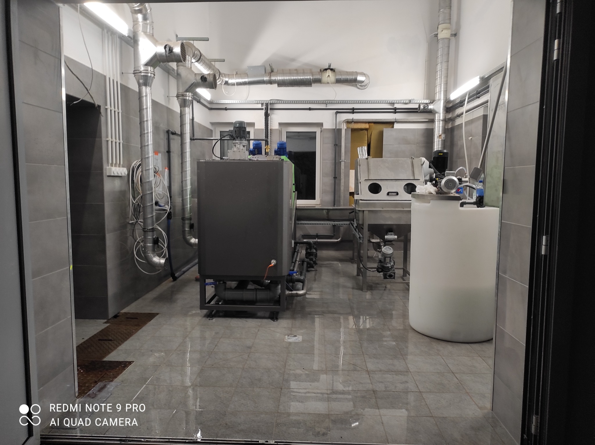 Wnętrze przemysłowe z instalacją rur wentylacyjnych pod sufitem, zbiornikiem i sprzętem do obróbki metali, widoczne płytki podłogowe