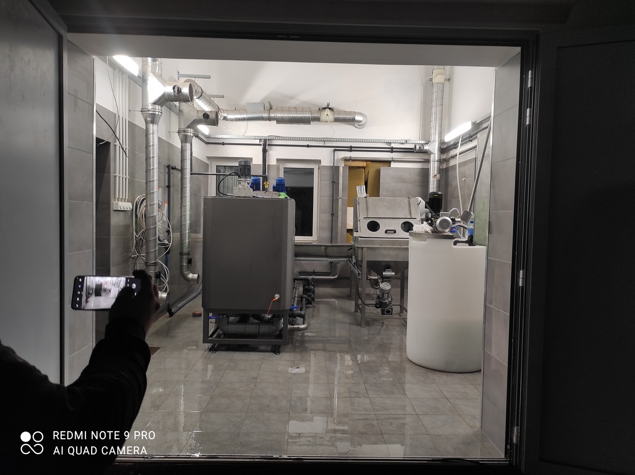 Wnętrze hali produkcyjnej z instalacją przemysłową, widok przez otwarte drzwi, osoba fotografuje wnętrze telefonem.