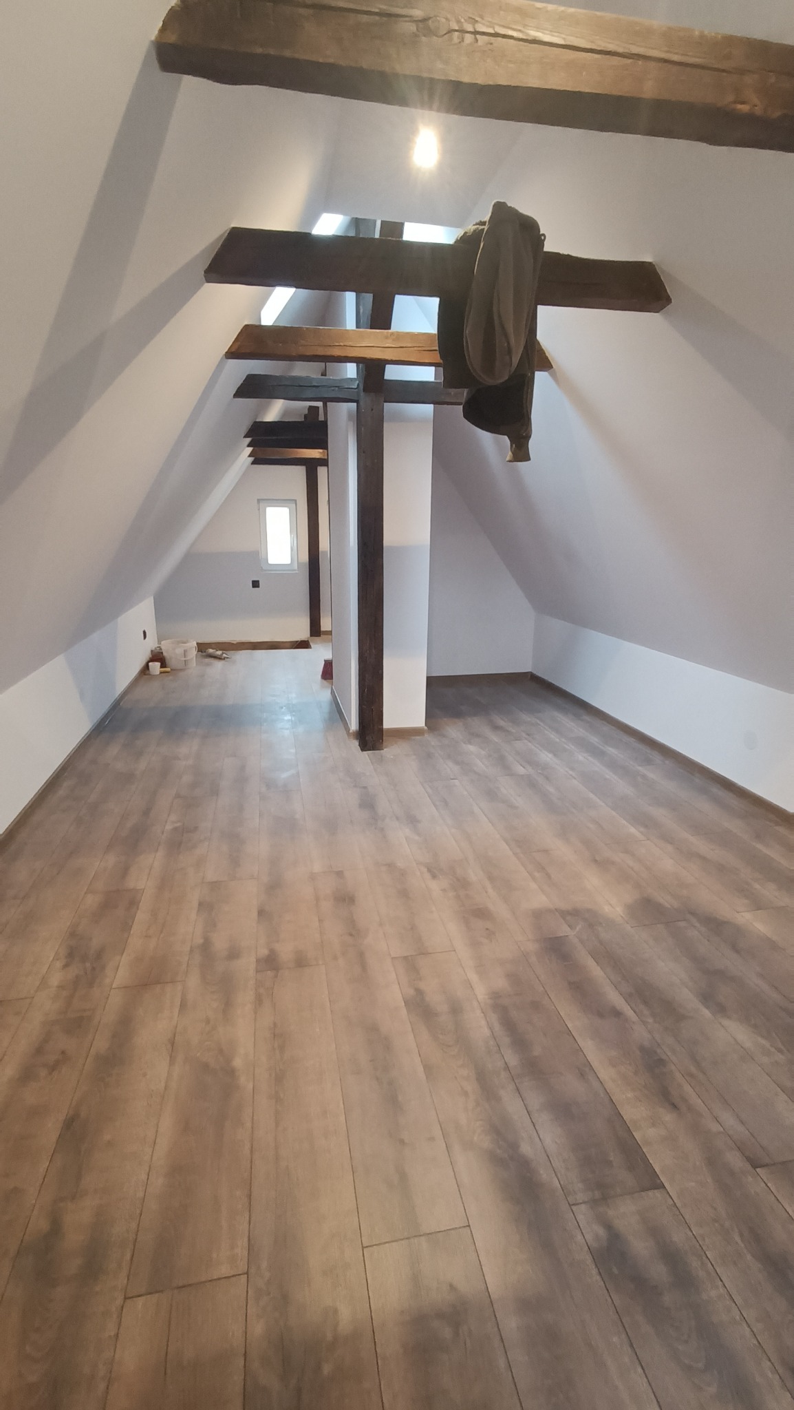 Poddasze po remoncie z nową podłogą z paneli imitujących drewno, widoczne drewniane belki stropowe i ubranie wiszące na jednej z nich.