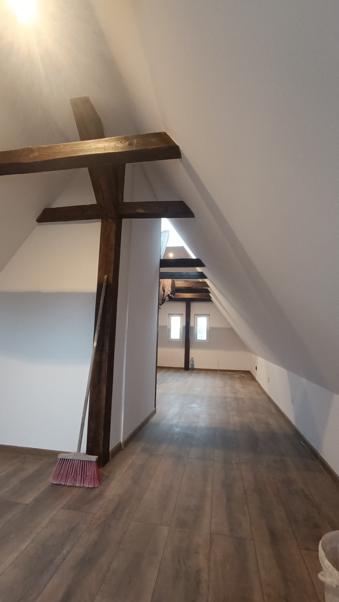 Poddasze po remoncie z drewnianymi belkami, nową podłogą z paneli i świeżo pomalowanymi ścianami, widoczna miotła oparta o belkę.