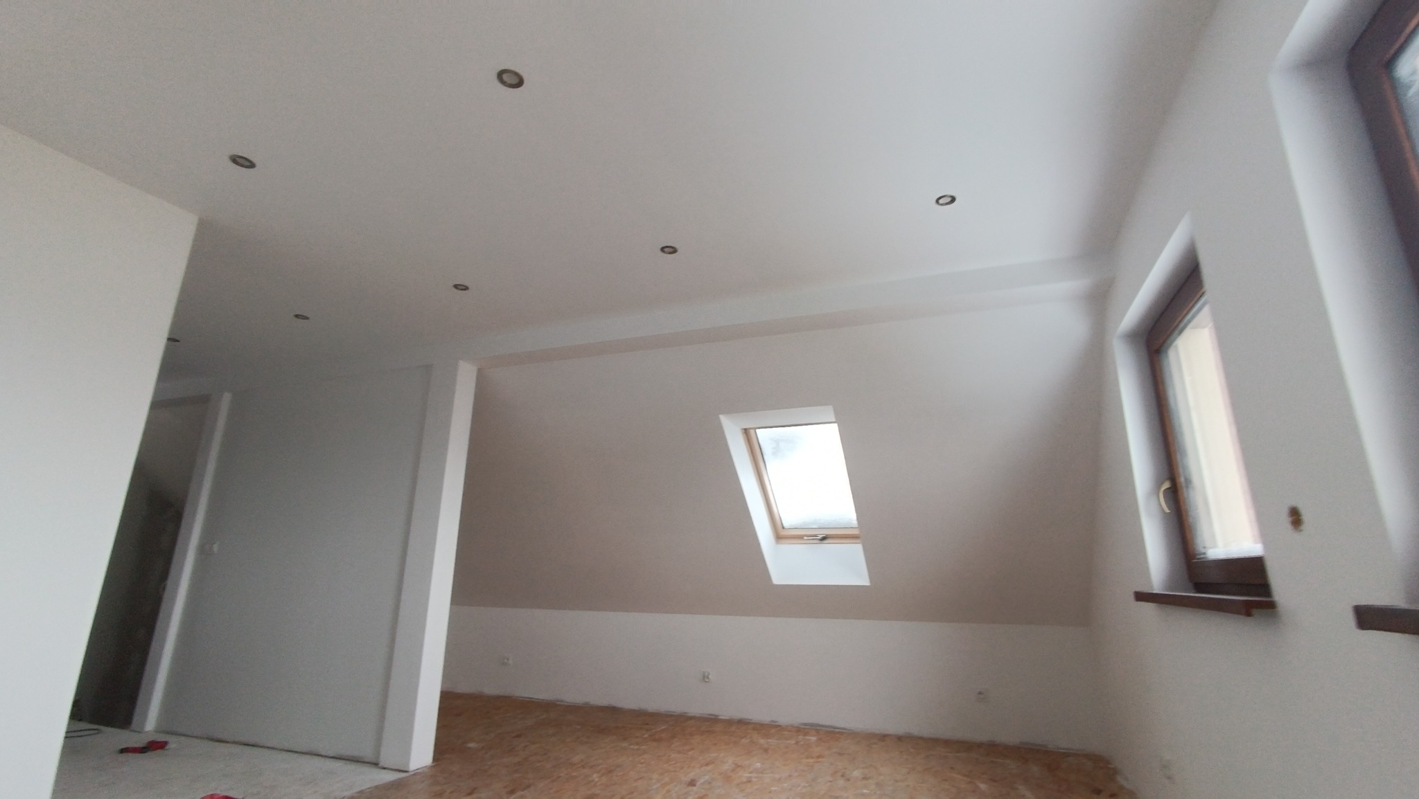 Poddasze po remoncie, świeżo pomalowane na biało, z widocznym oknem dachowym i zamontowanymi punktowymi lampami sufitowymi, podłoga przygotowana do dalszych prac.