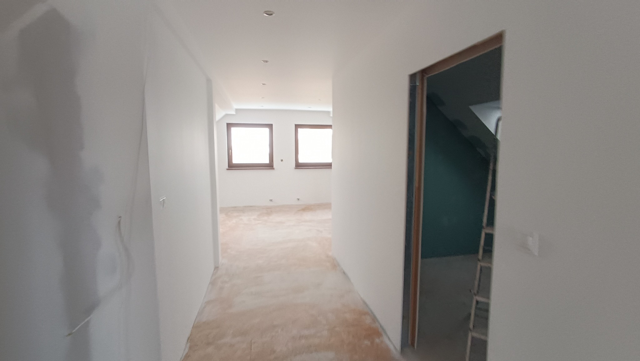 Wnętrze w trakcie remontu: korytarz z widokiem na pokój z dwoma oknami, ściany pomalowane na biało, podłoga przygotowana do układania paneli, widoczna drabina w przejściu.