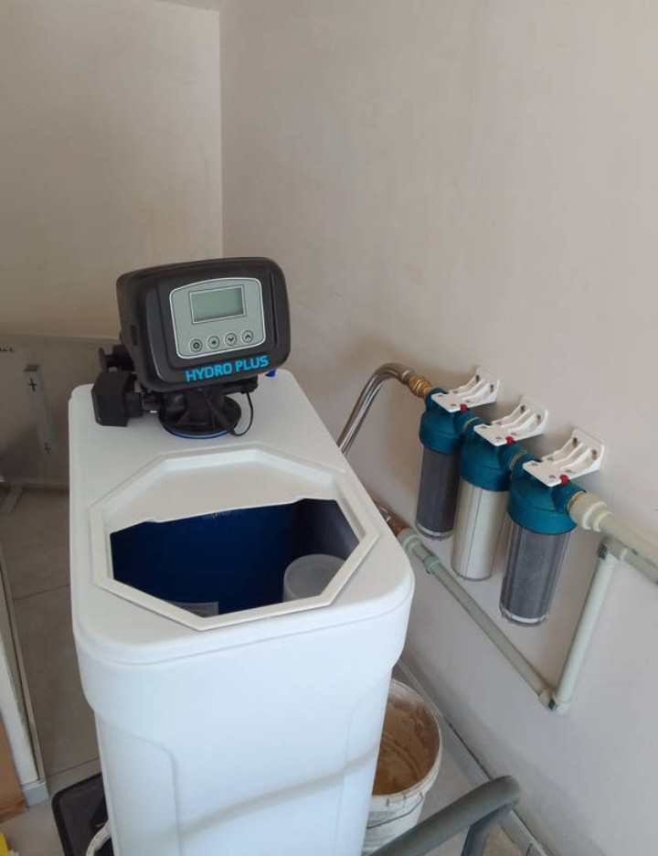 System filtracji wody Hydro Plus z trzema filtrami zainstalowany przy ścianie, widoczny pojemnik z otwartą pokrywą i elektroniczny panel sterowania.