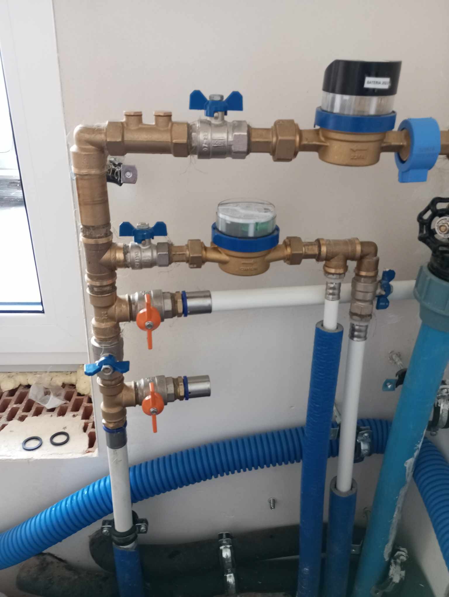 Kompleksowy system rur wodnych z zaworami, licznikiem i izolacją, zamontowany przy oknie z widocznymi uszczelkami i pianką montażową.