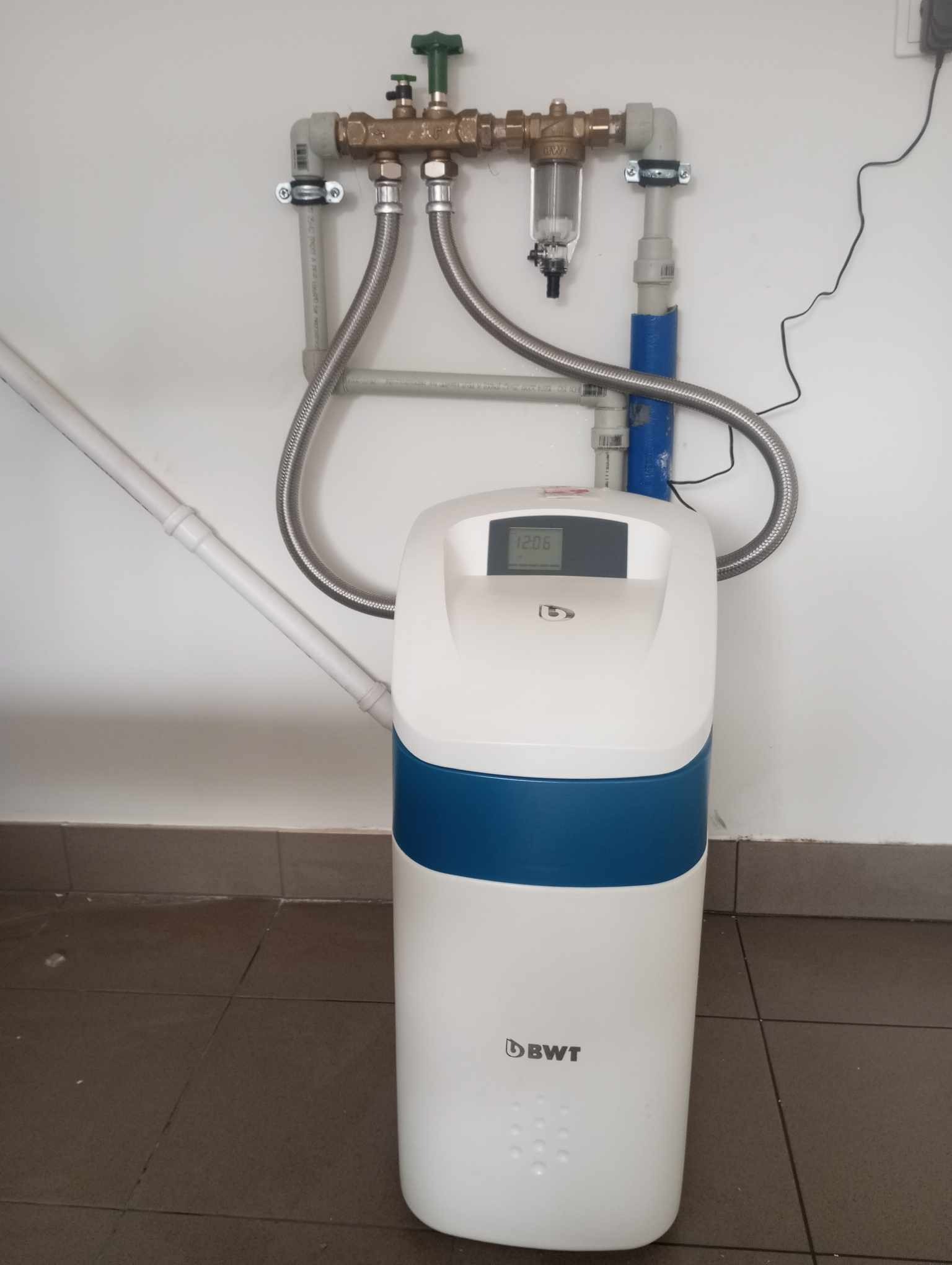 Stacja uzdatniania wody BWT z podłączeniami hydraulicznymi i filtrem wstępnym, zamontowana przy ścianie na brązowych płytkach podłogowych.