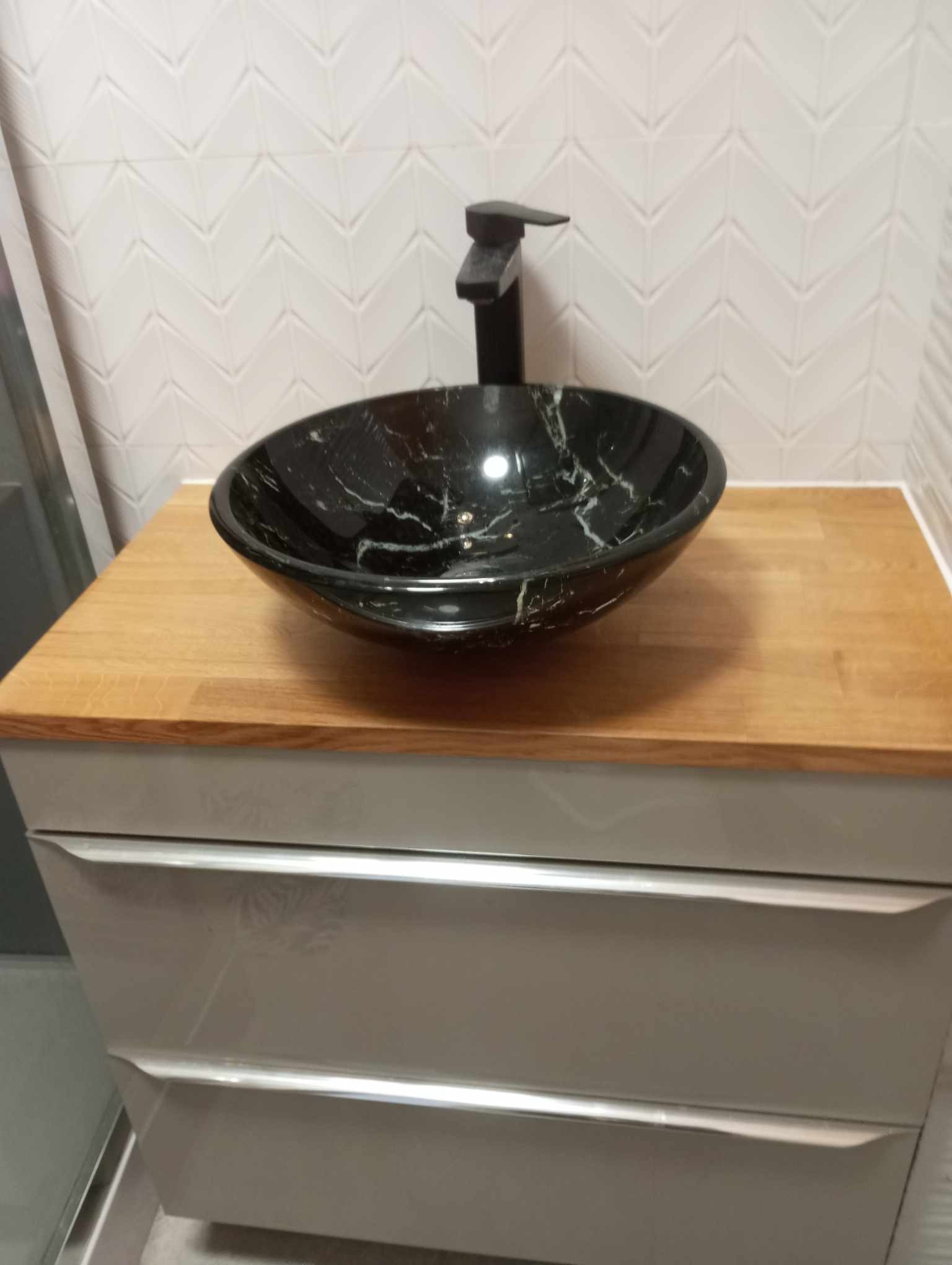 Czarna, marmurkowa umywalka nablatowa z czarną baterią, zamontowana na drewnianym blacie nad szarą szafką z trzema szufladami, na tle białych płytek ułożonych w jodełkę.