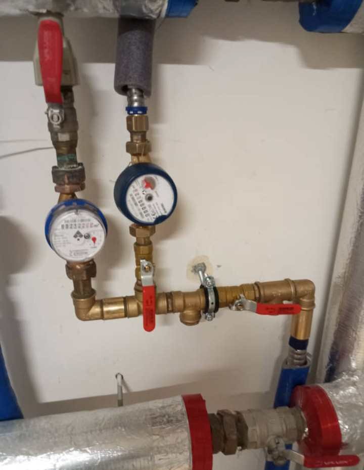 Fragment instalacji wodnej z licznymi zaworami z czerwonymi uchwytami, dwoma wodomierzami z niebieskimi obudowami i rurami miedzianymi oraz izolowanymi rurami na tle białej ściany.