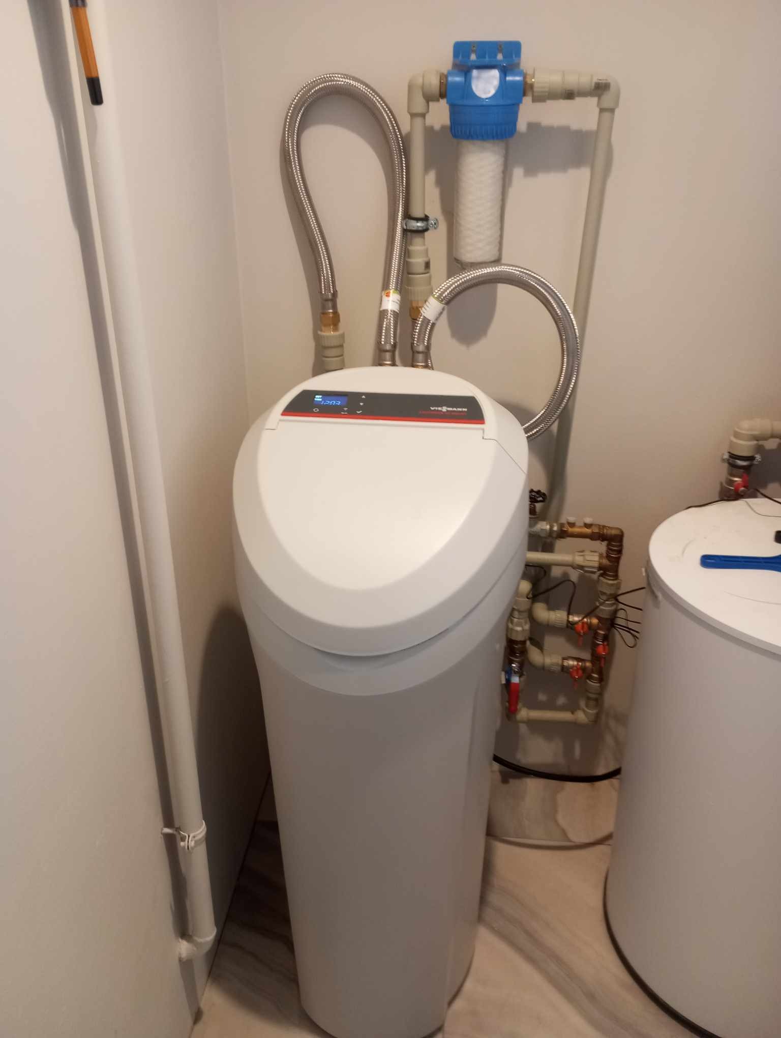 Nowoczesny, biały zmiękczacz wody Viessmann z wyświetlaczem, podłączony elastycznymi przewodami do instalacji z filtrem narurowym, w pomieszczeniu technicznym.