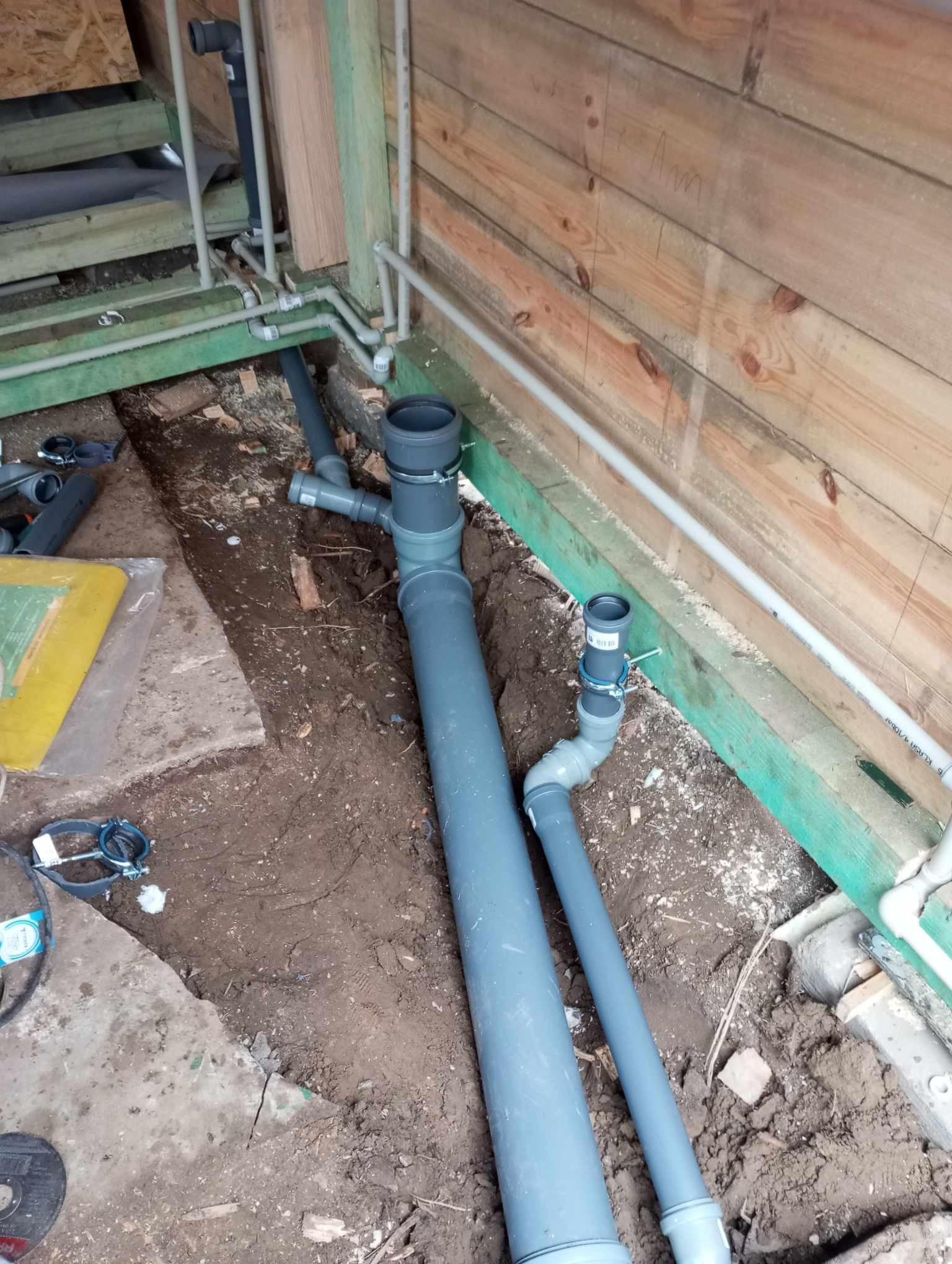 Szare rury kanalizacyjne z łącznikami, częściowo zakopane w wykopie w surowym stanie, widoczne drewniane elementy konstrukcyjne budynku w tle.