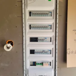 Biała skrzynka rozdzielcza z bezpiecznikami zamontowana na szarej ścianie w trakcie prac remontowych, widoczne przewody w pomarańczowej puszce instalacyjnej i odręczne notatki na kartonie.