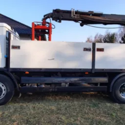 Biała ciężarówka z dźwigiem hydraulicznym na tle domu jednorodzinnego i drzew