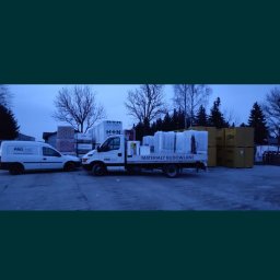 PRO-MAT Mateusz Taciak - Dwa samochody dostawcze firmy PRO-MAT na placu z materiałami budowlanymi, w tle widoczne palety z bloczkami H+H i YTONG oraz białe paczki z folią.