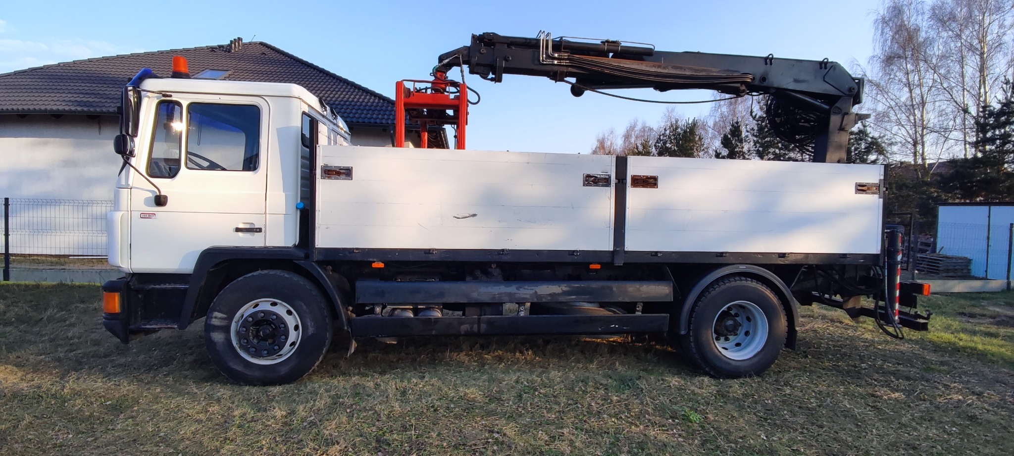 Biała ciężarówka z dźwigiem hydraulicznym na tle domu jednorodzinnego i drzew