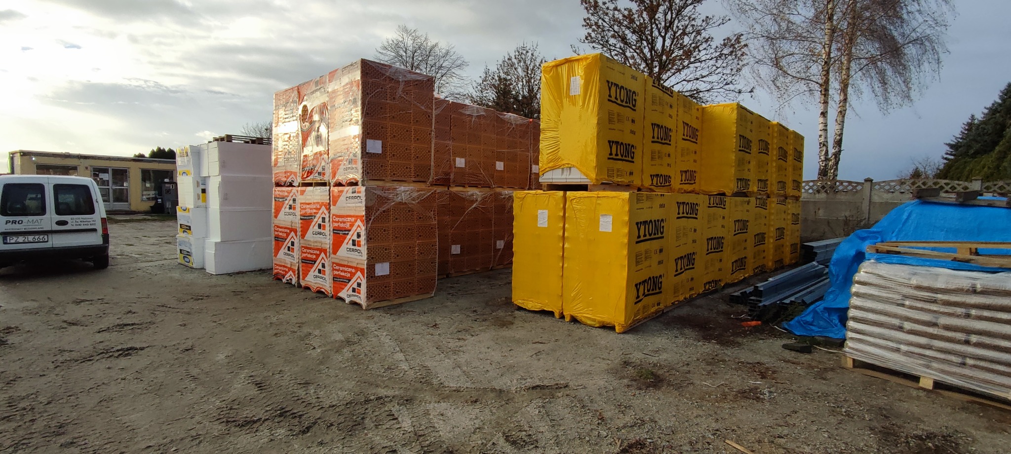 Skladowane materiały budowlane na placu: białe płyty izolacyjne, palety z cegłą ceramiczną Cerpol oraz żółte paczki Ytong, widoczny budynek w tle i dostawczy samochód.