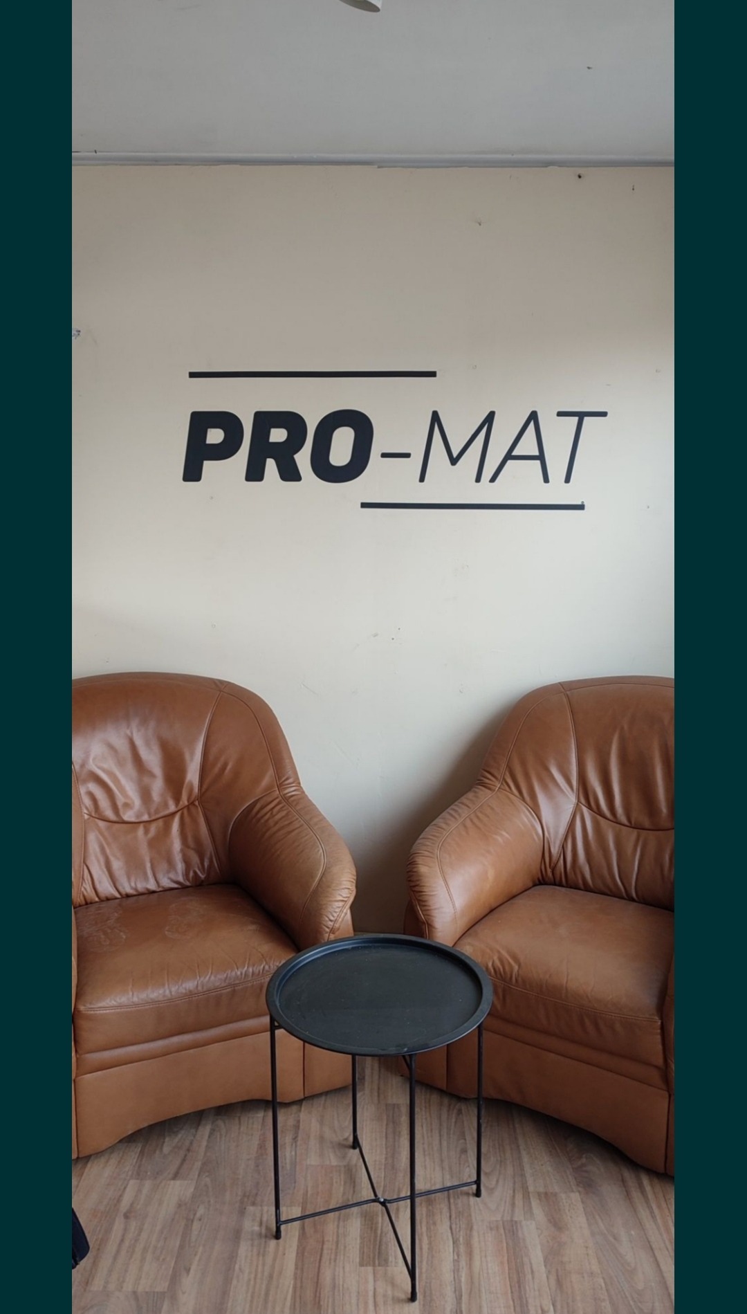 Dwa skórzane fotele w kolorze brązowym ustawione symetrycznie po bokach małego, czarnego stolika na metalowych nogach; na ścianie widnieje logo 'PRO-MAT'.