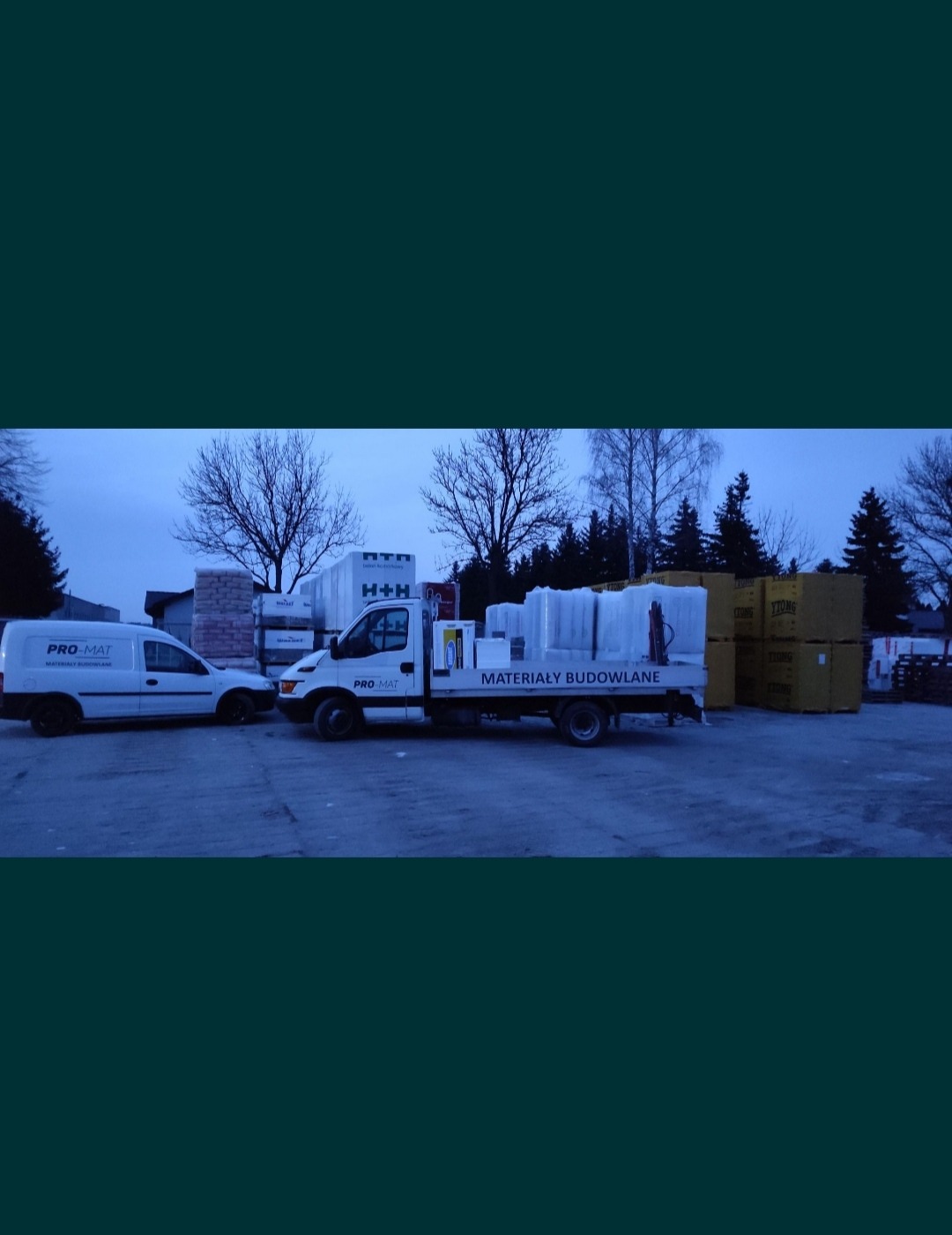 Dwa samochody dostawcze firmy PRO-MAT na placu z materiałami budowlanymi, w tle widoczne palety z bloczkami H+H i YTONG oraz białe paczki z folią.