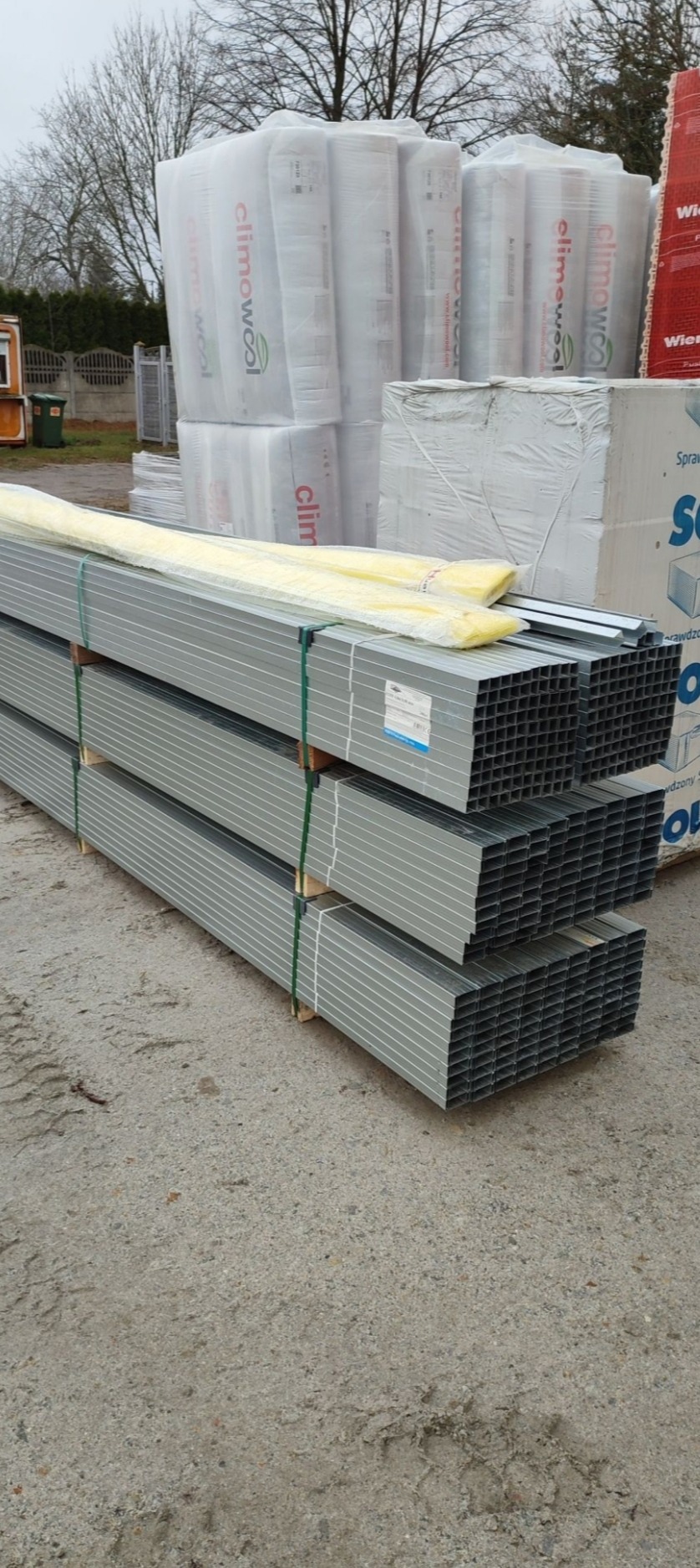 Stosy szarych, kwadratowych profili stalowych na paletach, owinięte zielonymi taśmami, z widocznymi etykietami, na tle zapakowanych paczek wełny mineralnej i styropianu.