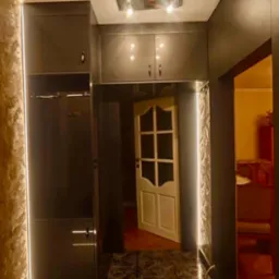 Nowoczesna szafa wnękowa z oświetleniem LED w przedpokoju, z połyskującymi frontami i marmurową podłogą. Widoczny fragment otwartych drzwi do innego pomieszczenia.