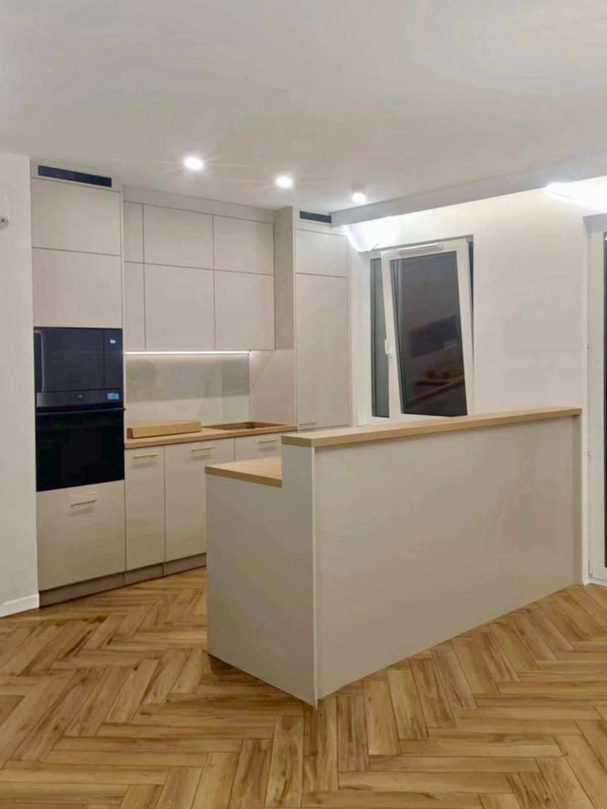 Jasna kuchnia z drewnianym parkietem w jodełkę. Zabudowa kuchenna z wyspą, piekarnik w zabudowie. Okno uchylone, w tle schody.