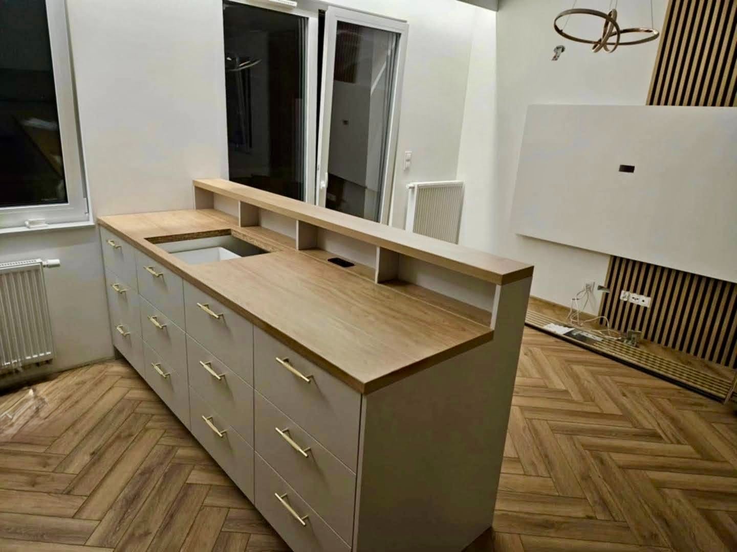 Nowoczesna zabudowa kuchenna z drewnianym blatem i białymi frontami, złote uchwyty, parkiet w jodełkę, minimalistyczne oświetlenie i dekoracyjna ściana z lameli.