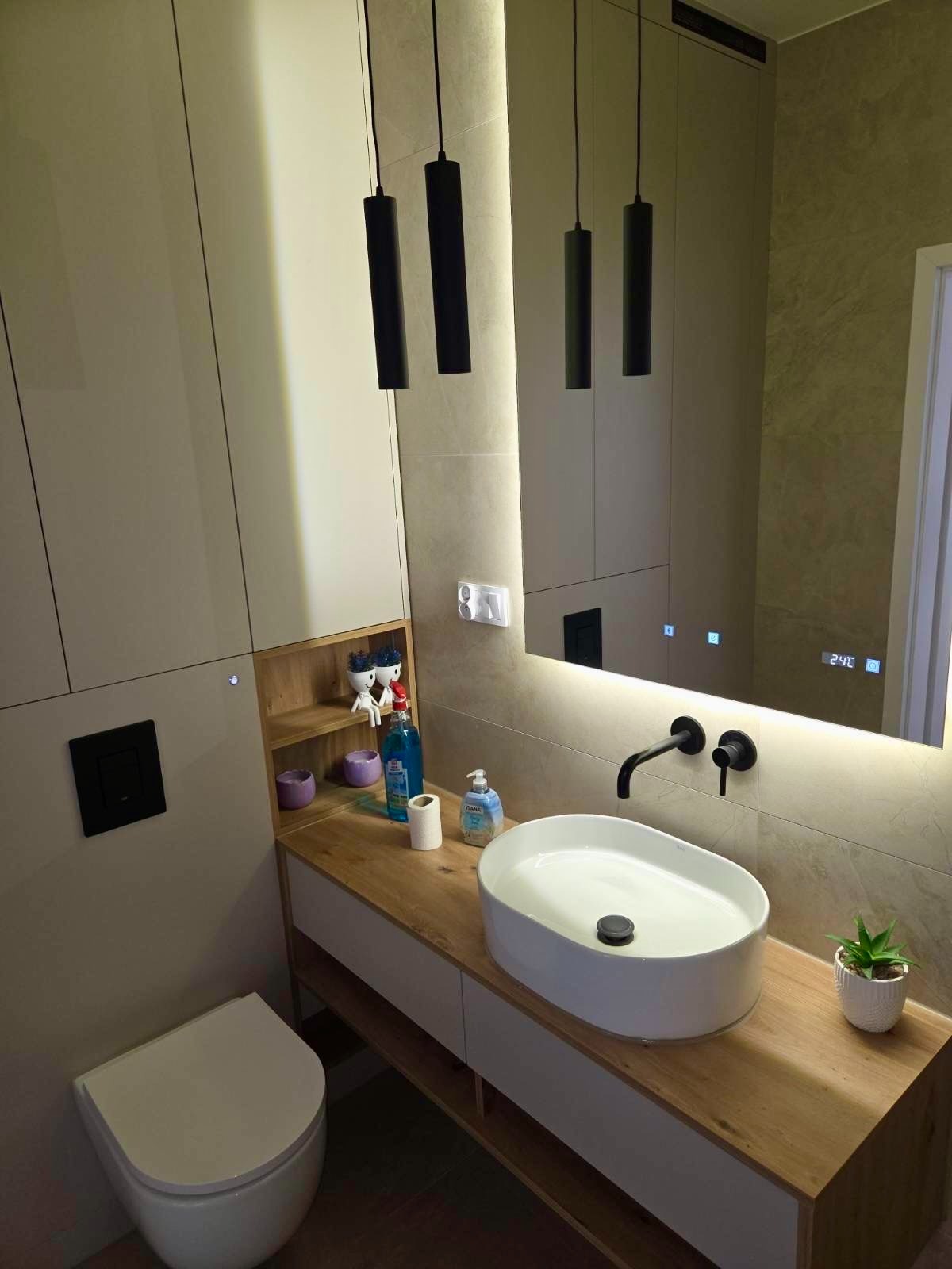 Nowoczesna łazienka z szafką pod umywalką z drewnianym blatem, podświetlanym lustrem i wiszącymi lampami. Uwagę zwraca minimalistyczny design i stonowana kolorystyka.