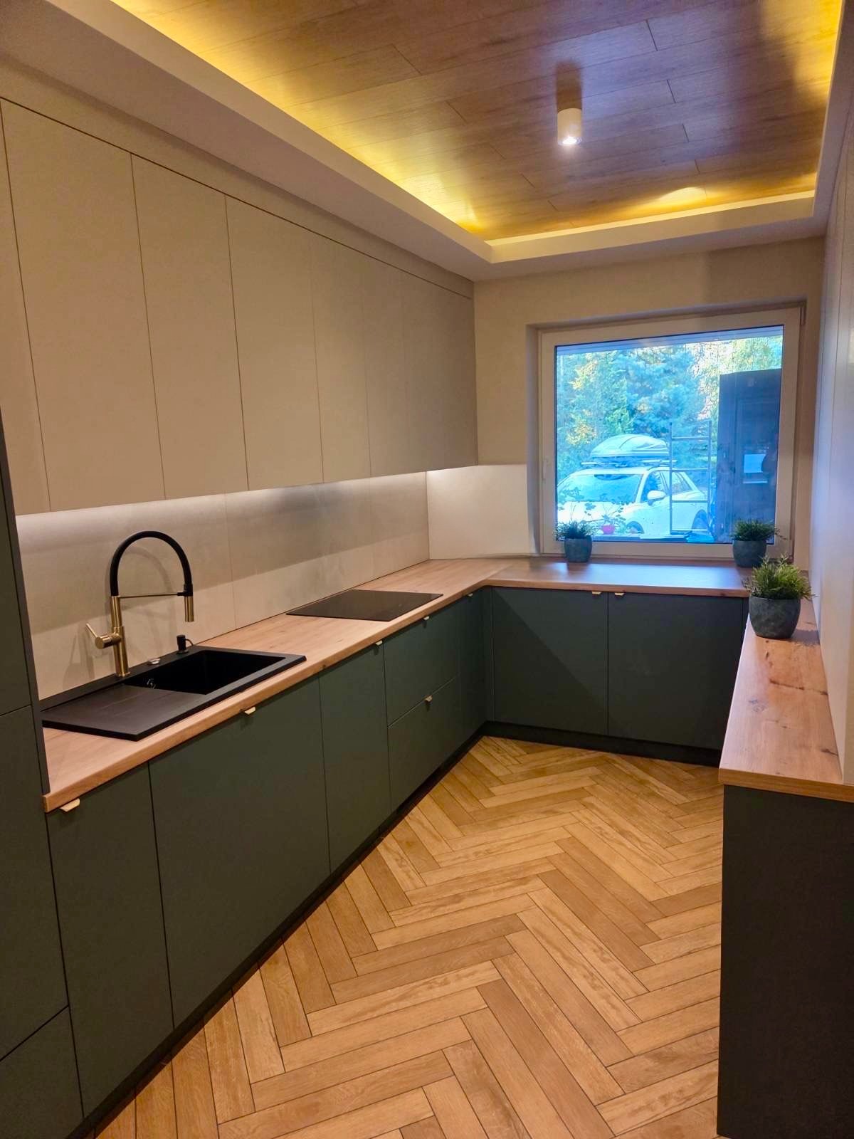 Nowoczesna kuchnia w odcieniach beżu i zieleni z drewnianym blatem i parkietem w jodełkę. Uwagę przykuwa złota bateria i oświetlenie LED podszafkowe.