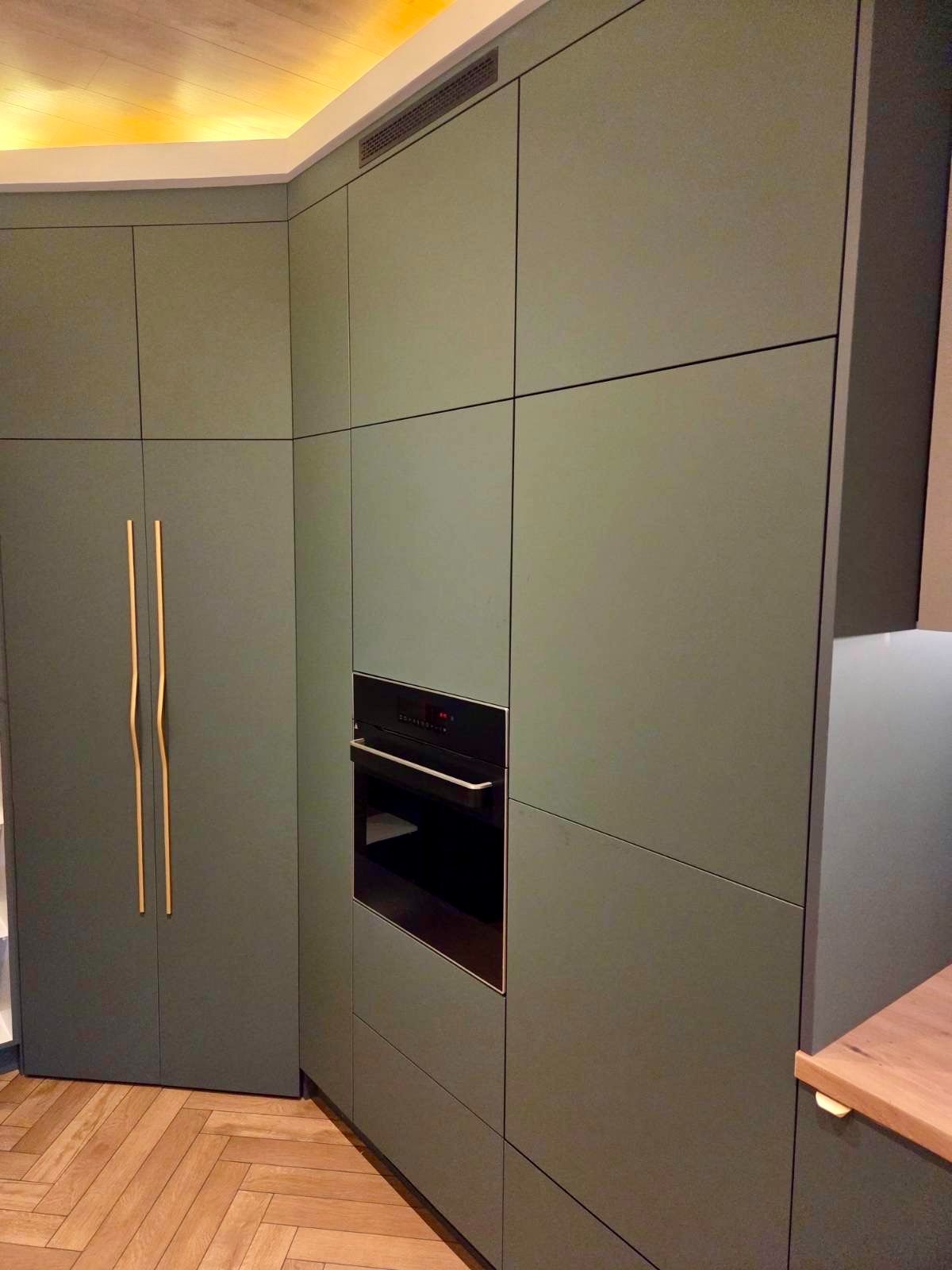 Nowoczesna kuchnia w odcieniach zieleni z minimalistycznymi, złotymi uchwytami i wbudowanym piekarnikiem. Parkiet w jodełkę na podłodze.