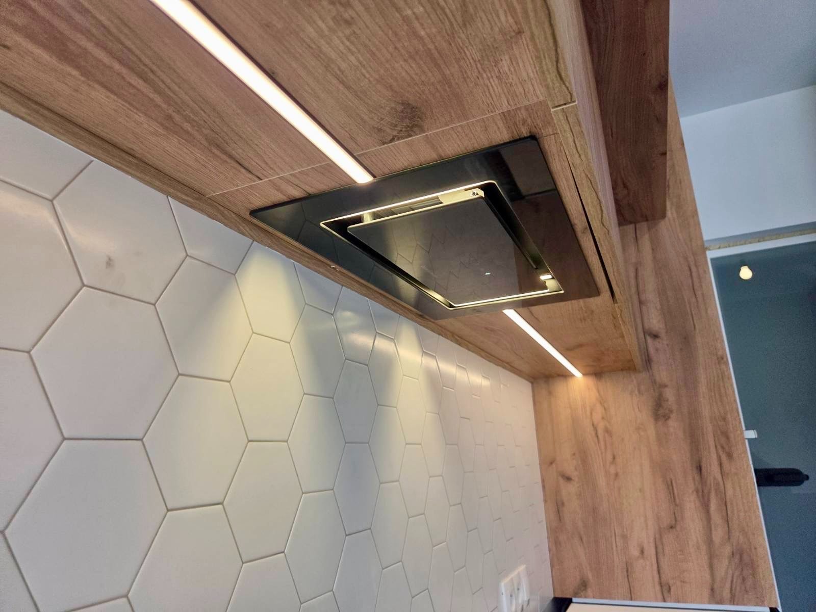 Nowoczesny okap podszafkowy z oświetleniem LED, wkomponowany w drewniane szafki kuchenne i białe heksagonalne płytki. Ujęcie z dołu, podkreślające detale wykończenia.