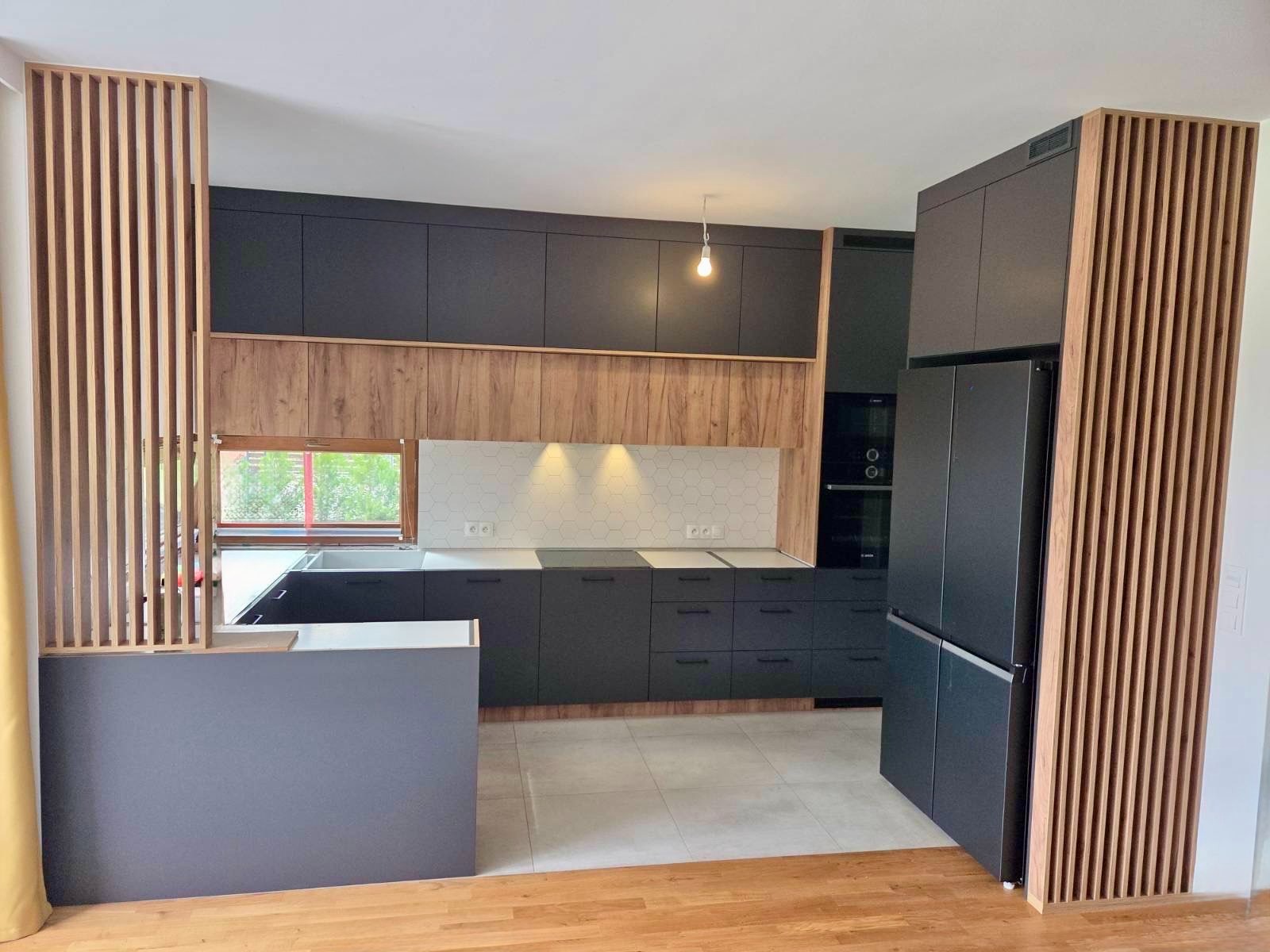 Nowoczesna kuchnia z ciemnymi szafkami, drewnianymi akcentami i lamelami. Widoczny fragment wyspy, płytki heksagonalne nad blatem i sprzęt AGD w zabudowie.