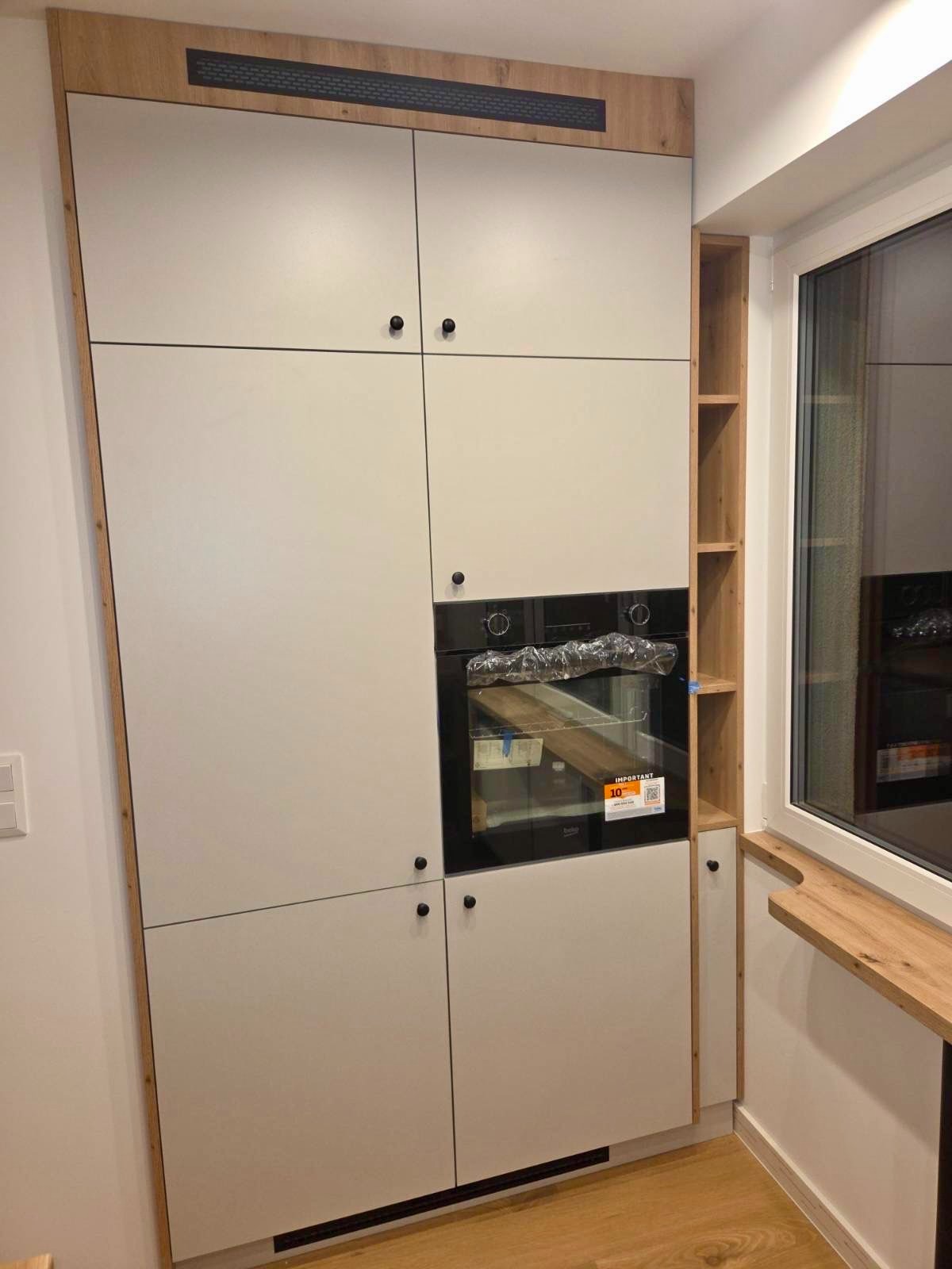 Jasna zabudowa kuchenna z piekarnikiem i drewnianymi akcentami, dopasowana do okna z półkami. Minimalistyczny design, czarne okrągłe uchwyty.