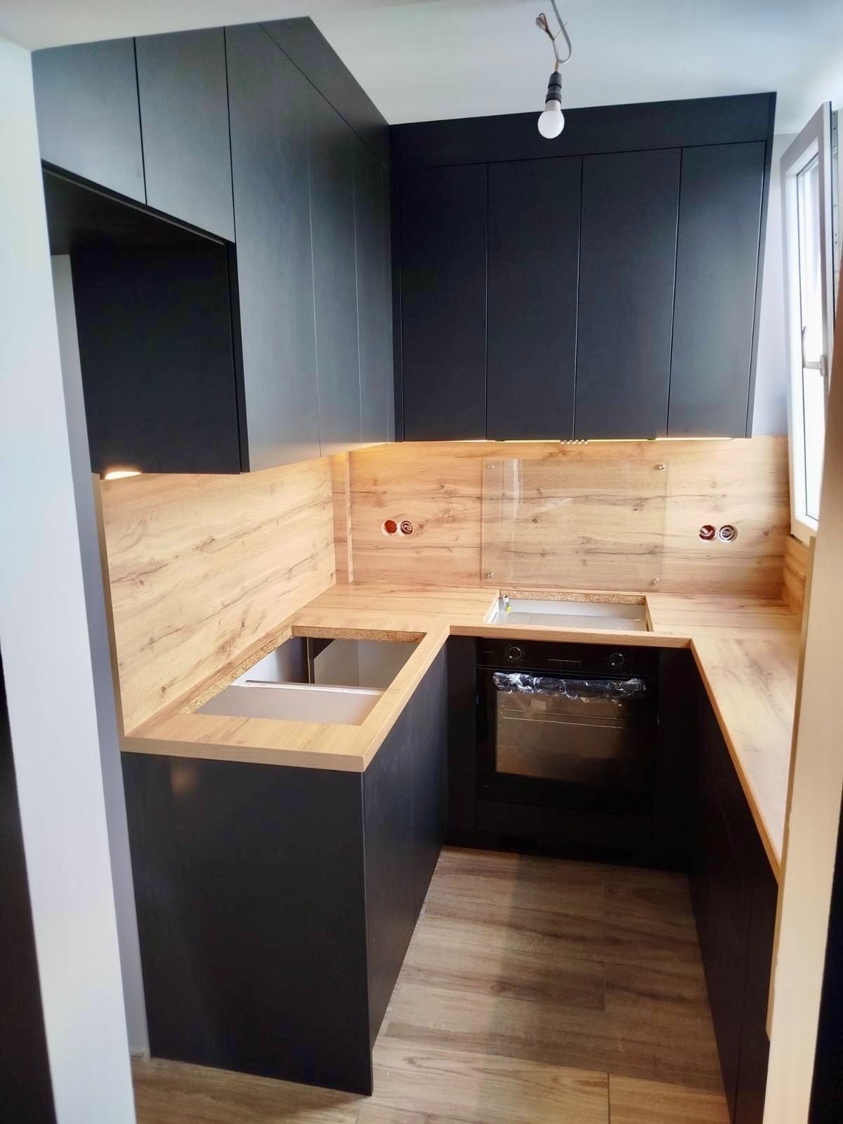 Nowoczesna kuchnia w kształcie litery U z czarnymi szafkami i drewnianym blatem. Widoczne gniazdka elektryczne i oświetlenie podszafkowe. Ujęcie z perspektywy wejścia.