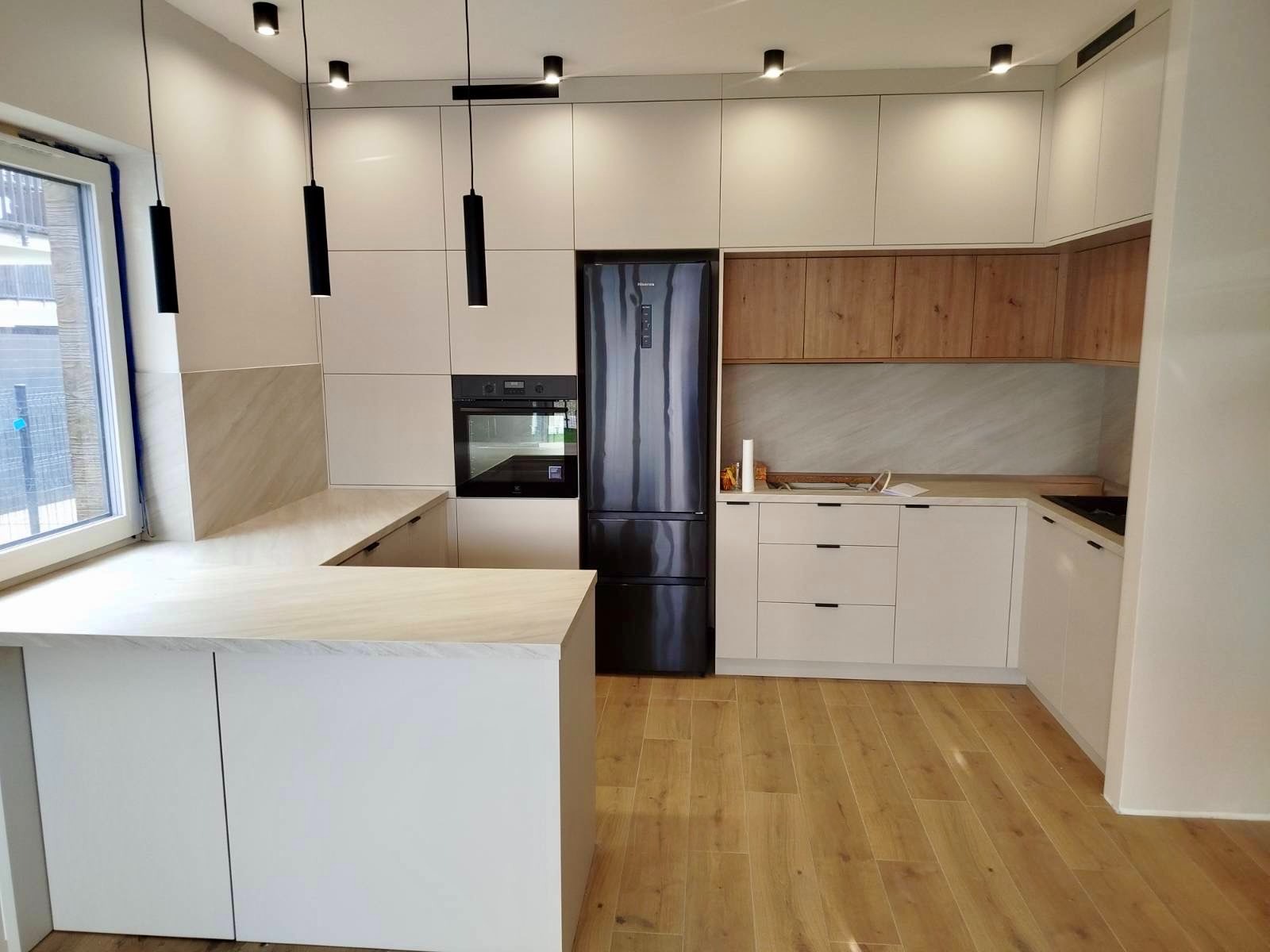 Nowoczesna kuchnia z wyspą: białe fronty, drewniane akcenty, czarny sprzęt AGD i podłużne lampy wiszące. Jasna podłoga i okno z widokiem na zewnątrz.