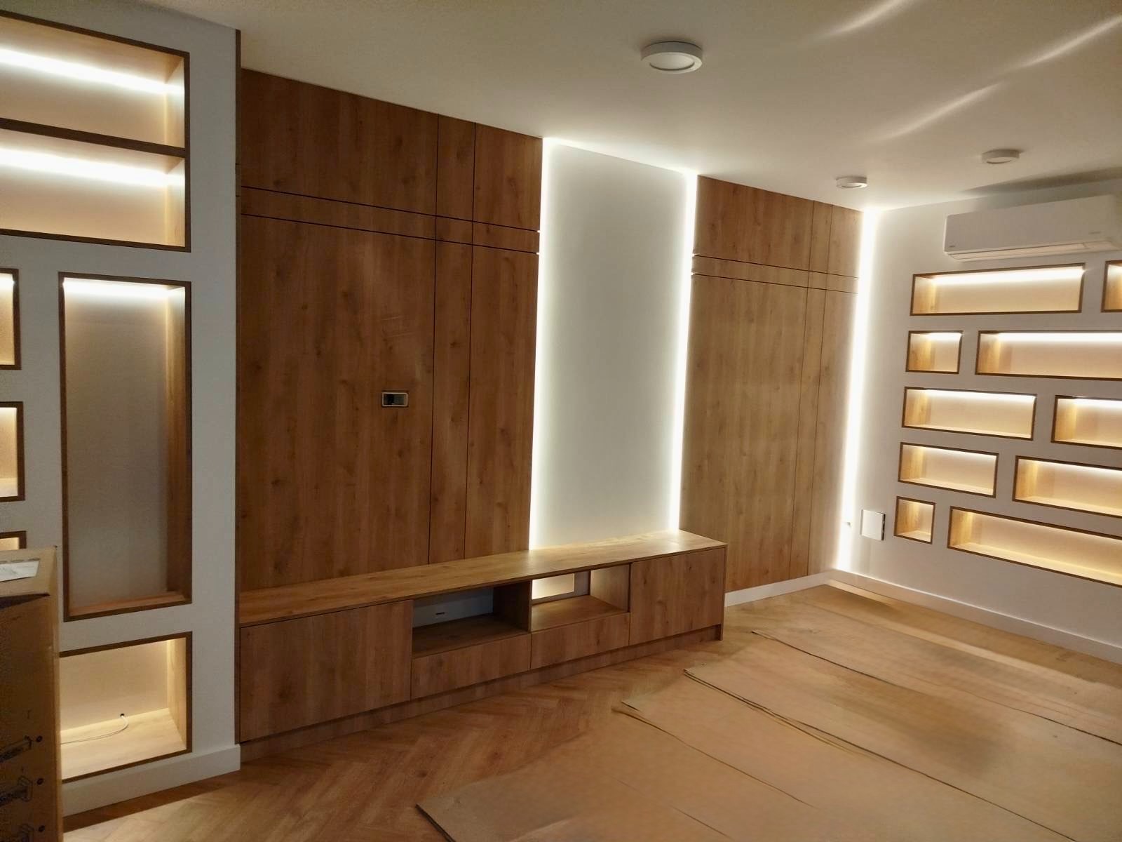 Drewniana zabudowa ściany z szafami i półkami podświetlonymi LED, z widocznym parkietem w jodełkę i klimatyzacją. Minimalistyczny design z ciepłym oświetleniem.