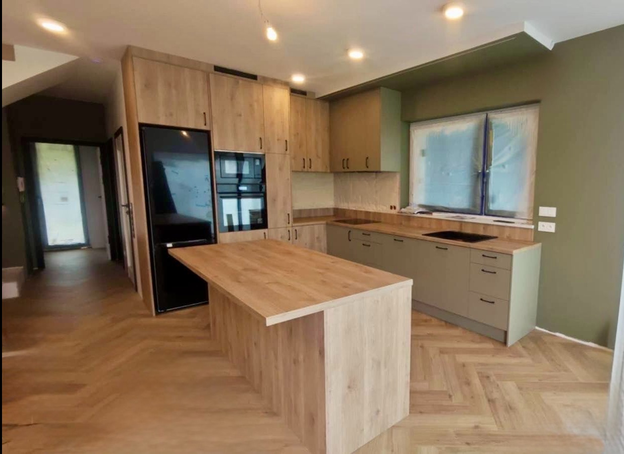 Jasna kuchnia z drewnianymi frontami i wyspą, czarny sprzęt AGD, parkiet w jodełkę. Ściany w odcieniach zieleni. Okno zasłonięte folią malarską.