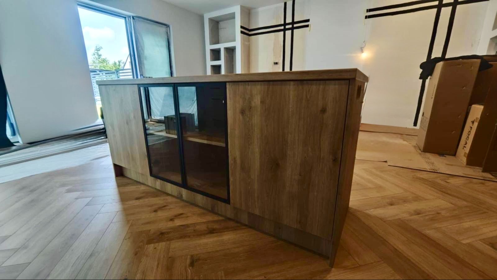 Drewniana szafka z przeszklonymi drzwiczkami na tle jasnego wnętrza w trakcie wykańczania. Widoczna podłoga ułożona w jodełkę i okno przesuwne.