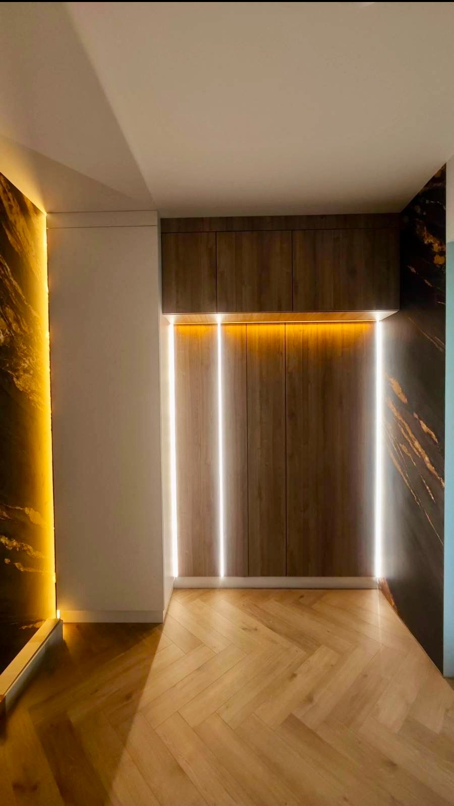 Nowoczesna szafa wnękowa z oświetleniem LED, drewniane fronty, parkiet w jodełkę, marmurowa ściana z podświetleniem. Minimalistyczny design, ciepłe światło.