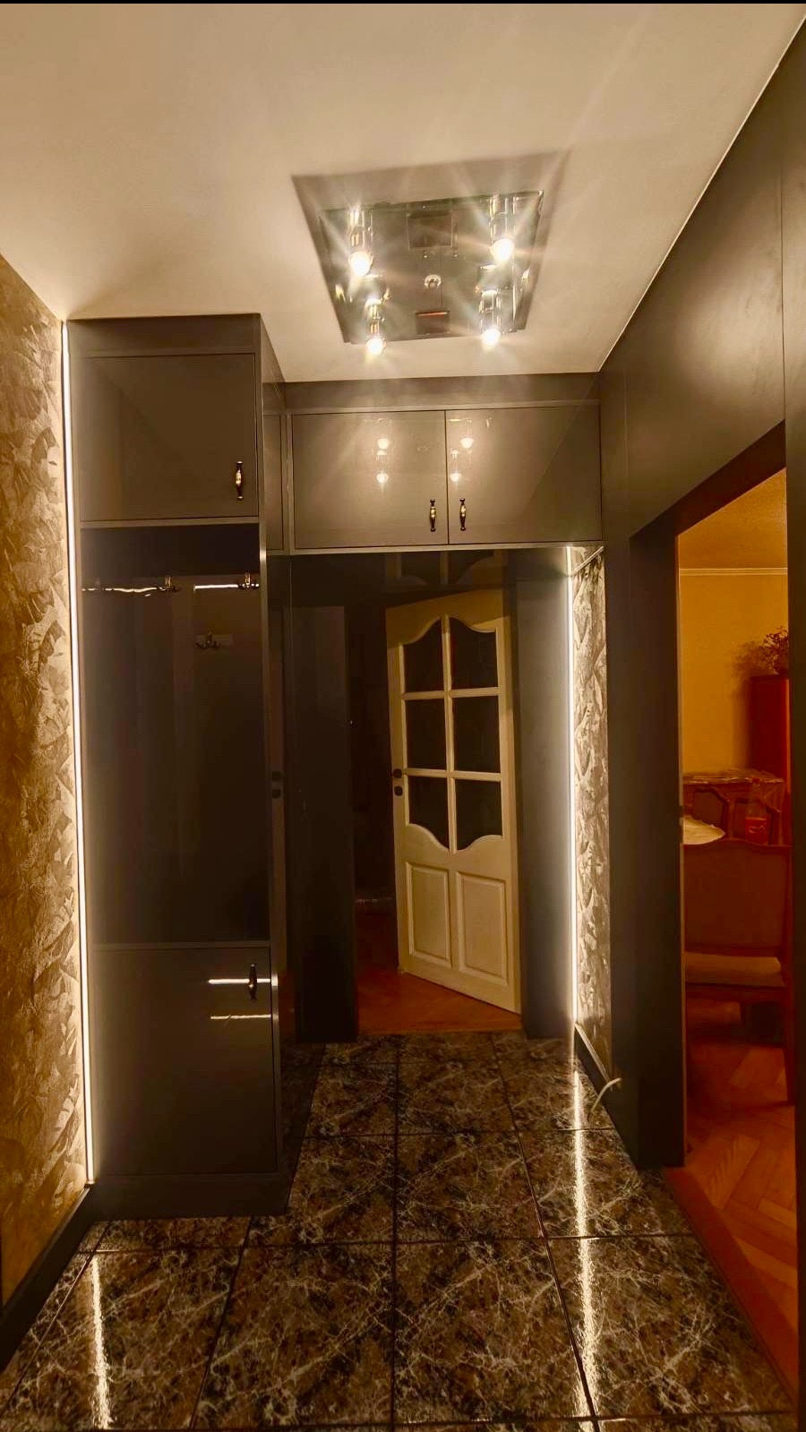 Nowoczesna szafa wnękowa z oświetleniem LED w przedpokoju, z połyskującymi frontami i marmurową podłogą. Widoczny fragment otwartych drzwi do innego pomieszczenia.