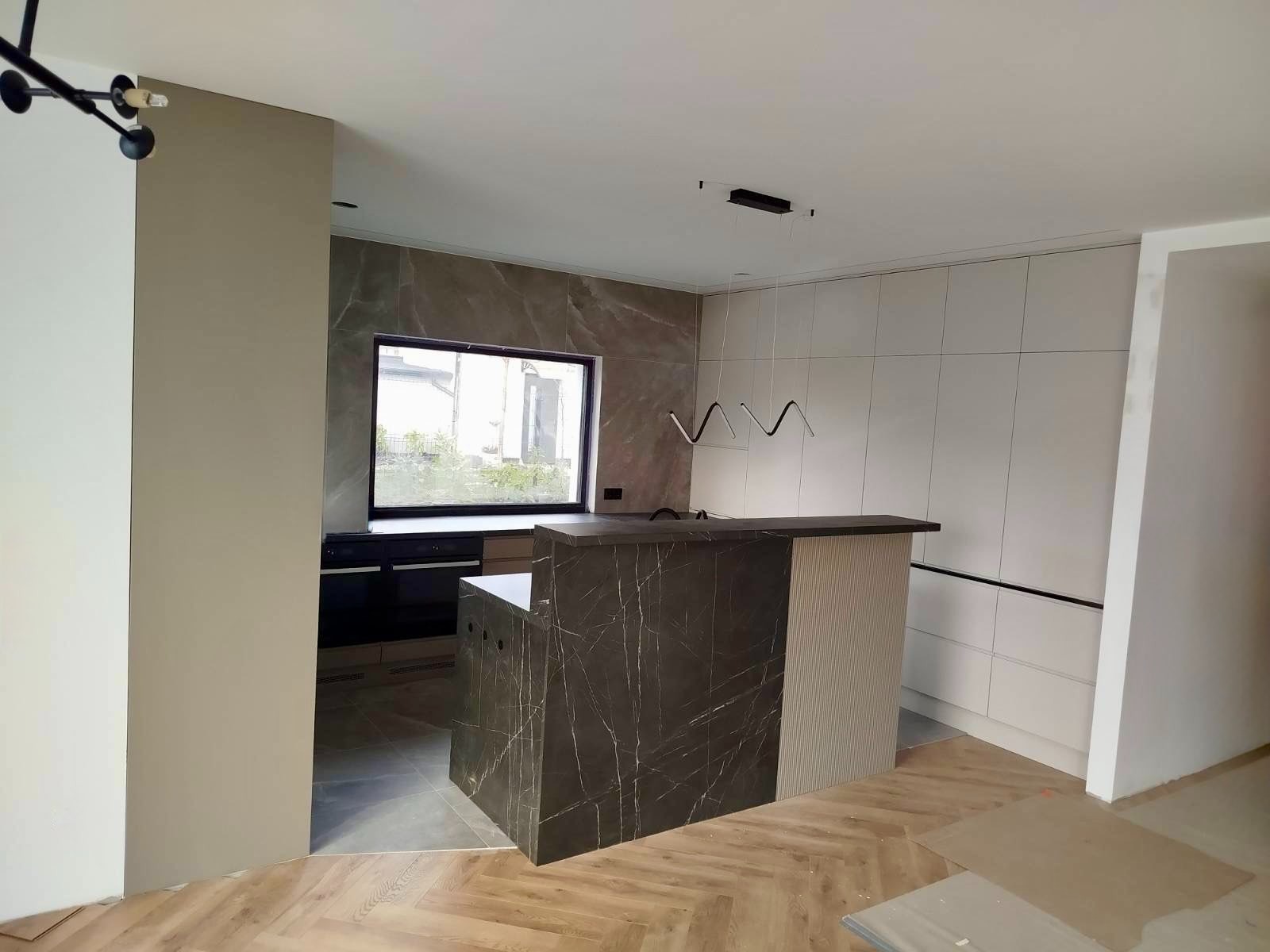 Nowoczesna kuchnia z wyspą wykończoną marmurem i ryflowanym panelem, minimalistyczne oświetlenie, jasne szafki, okno z widokiem na zewnątrz, parkiet w jodełkę.