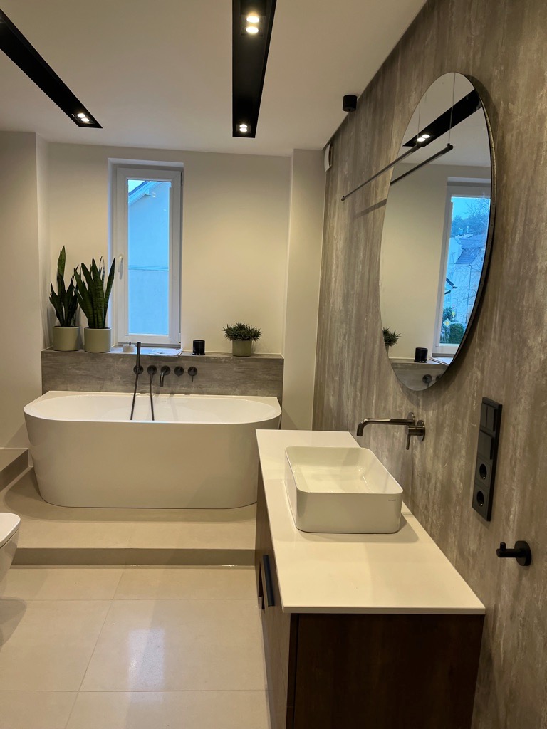 Nowoczesna łazienka z wolnostojącą wanną, umywalką nablatową na szafce z ciemnego drewna i dużym owalnym lustrem na ścianie o teksturze betonu. Czarna armatura i oświetlenie punktowe dodają elegancji.