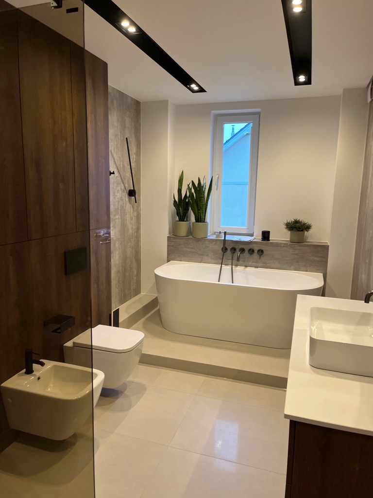 Nowoczesna łazienka z wolnostojącą wanną, wiszącą toaletą i bidetem, wykończona w beżach i brązach, z czarnymi akcentami w armaturze i oświetleniu sufitowym.
