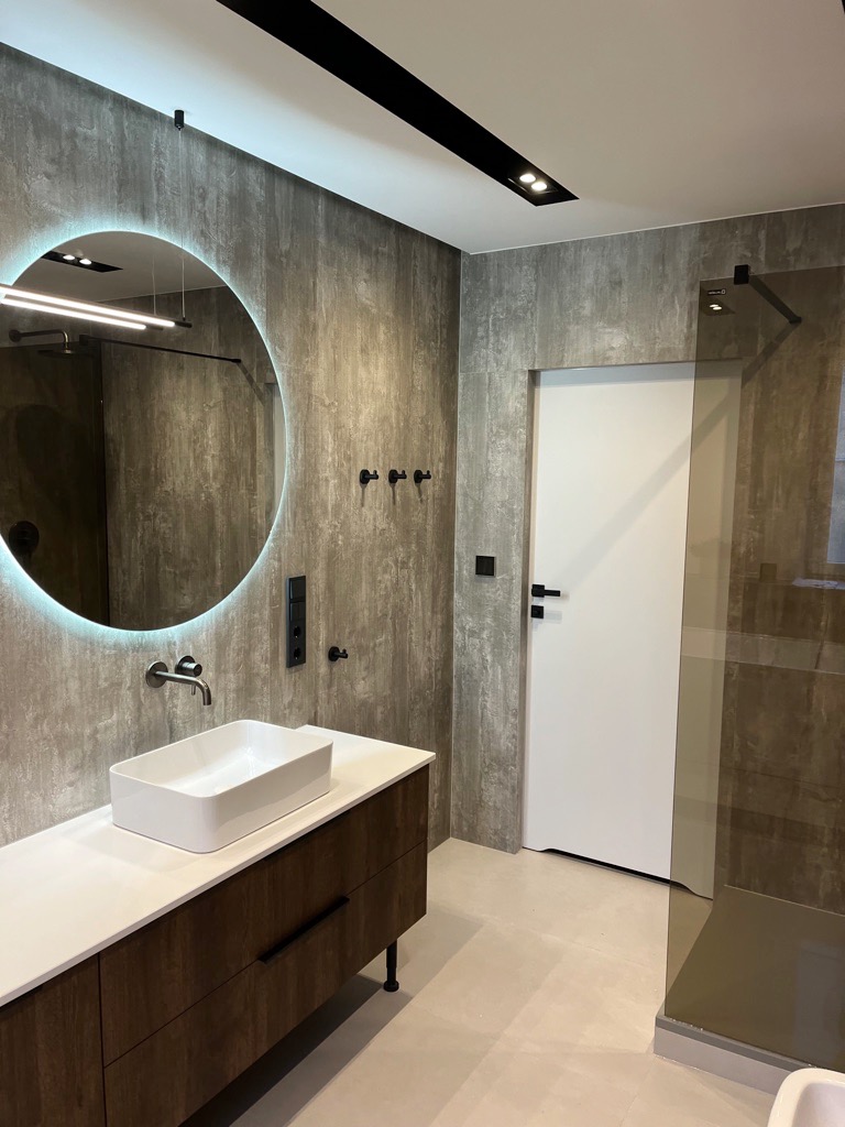 Nowoczesna łazienka z umywalką nablatową, okrągłym lustrem z podświetleniem LED, ścianami wykończonymi płytkami imitującymi beton i minimalistyczną kabiną prysznicową.