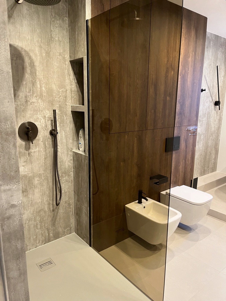 Nowoczesna łazienka z kabiną prysznicową typu walk-in, betonową ścianą, deszczownicą, czarną armaturą, zabudową z ciemnego drewna, wiszącym bidetem i toaletą.