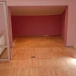 Wnętrze pokoju z ułożoną drewnianą podłogą z listew, różowe ściany i sufit z oświetleniem punktowym, widoczny fragment listwy przypodłogowej i mały czarny przedmiot na podłodze.